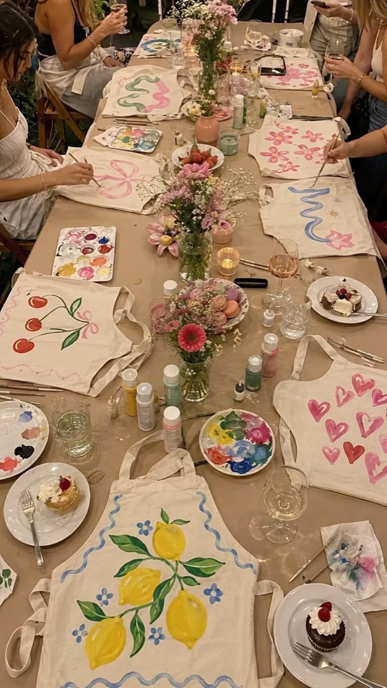 A long table of friends painting aprons with flowers and snacks