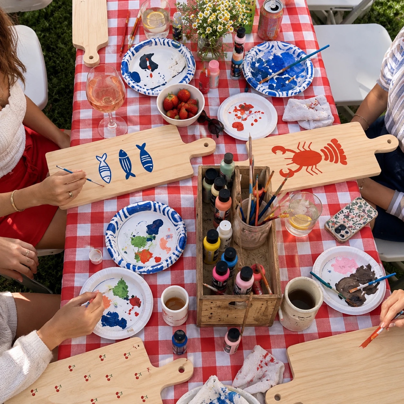 Friends painting wooden serving boards at a red gingham picnic table